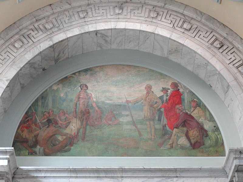 Two groups of people gather on a river bank. One group of American Indian people are wearing hair ornaments and deerskin clothing. The people among the group of white settlers are wearing colonial garb. The two central characters are likely Dragging Canoe and Daniel Boone.