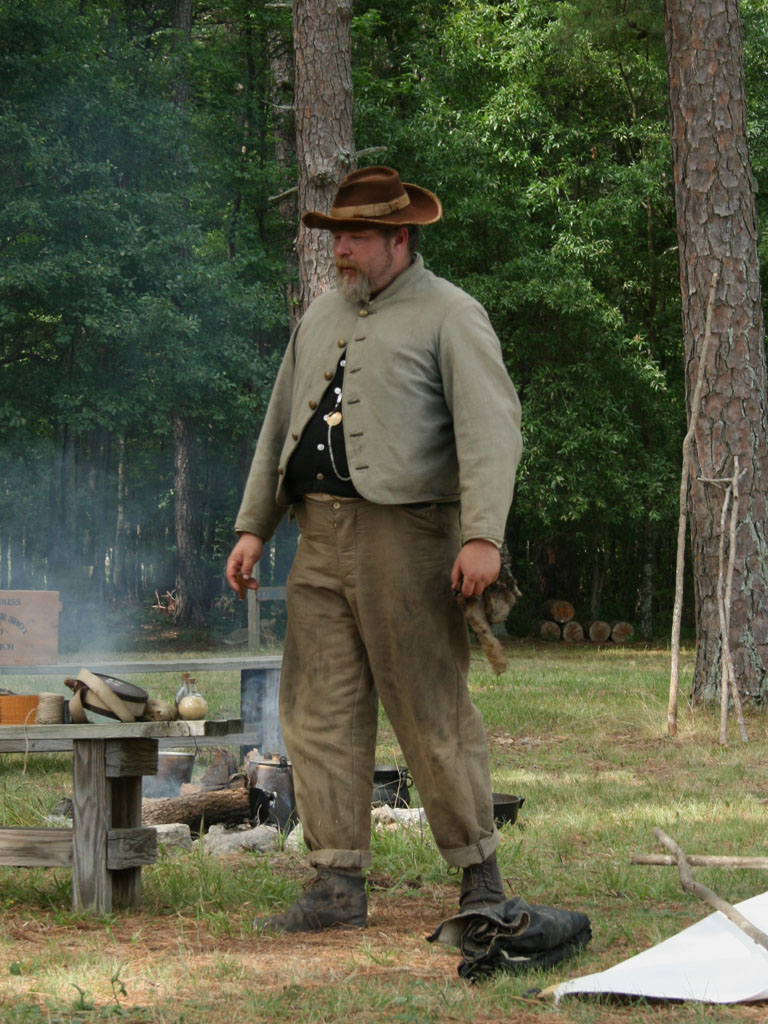 Image of a Confederate reenactor in a "butternut" uniform. He is wearing a brown hat, a grey jacket, and brown pants. He is in a wooded area. 