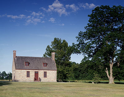 The brick house with a chimney, red door and windows it stands next to a large tree. 