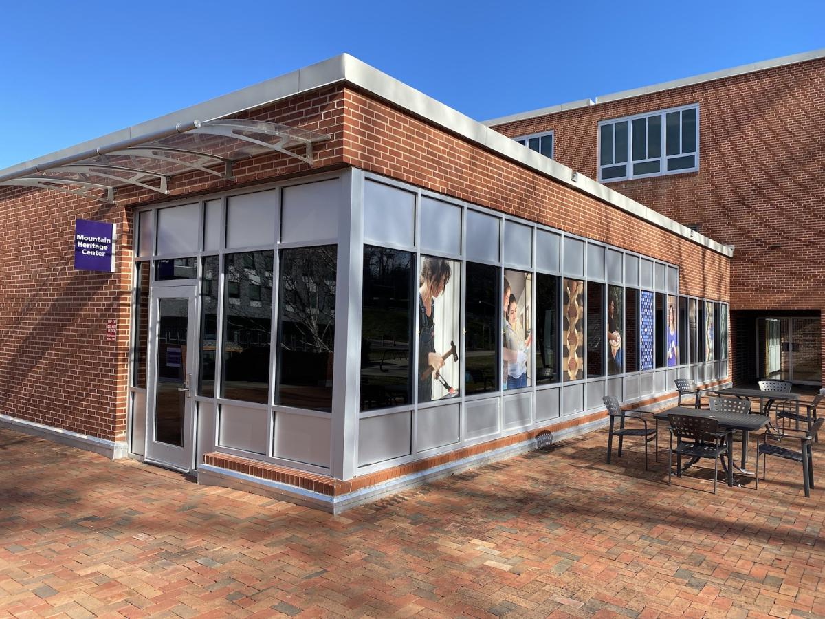 A brick building with glass windows at its front. A small purple sign on the left reads "Mountain Heritage Center."