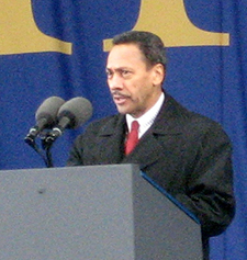 Melvin Watts. He is speaking at a lectern. He has a coat and tie on. He has short hair and a moustache.