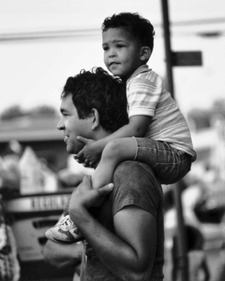 A man with dark hair stands in profile.  He is wearing a t-shirt and is smiling.  He has a small boy on his shoulders.  The boy also has dark hair and is wearing shorts and a short-sleeved collared shirt.  He is looking to the left of the camera and is smiling.