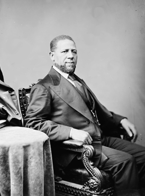 Revels seated. He is middle aged, with short hair and beard. He is wearing a suit. 