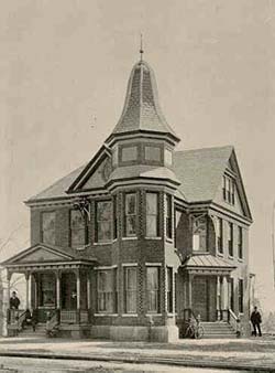 A Victorian style building.  On the left side of the image stairs lead up to a porch entry to the house.  A man rests against a porch post and another sits on the stairs. A turret is on the corner of the house which is in the center of this image. To the right is another set of stairs leading to another entry into the building.  A bicycle rests against the left handrail of the steps.  A man stands to the right of the steps.